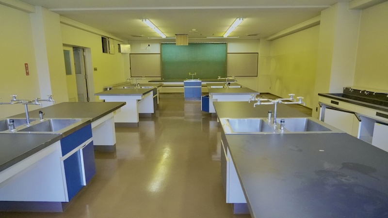 実験台と流し台が並ぶ化学実験室の室内写真。正面に黒板がある
