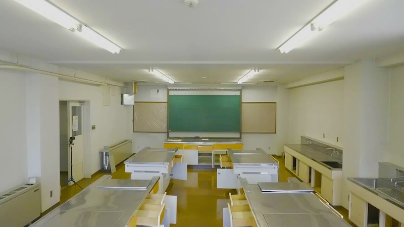 ステンレスの調理台とシンクが並ぶ学校の調理室。正面に黒板がある